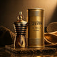 Jean Paul Gaultier Le Male Elixir perfume bottle and its cylindrical packaging box, both in a metallic gold finish, displayed on a dark stone pedestal with draped gold fabric in the background.