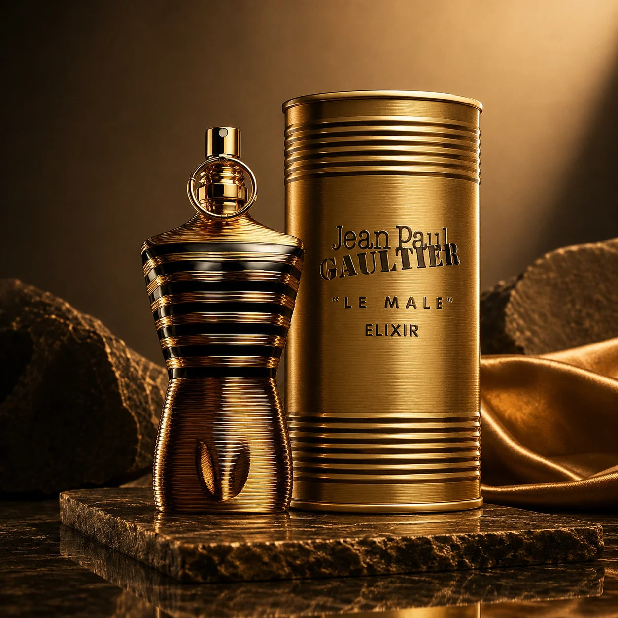 Jean Paul Gaultier Le Male Elixir perfume bottle and its cylindrical packaging box, both in a metallic gold finish, displayed on a dark stone pedestal with draped gold fabric in the background.