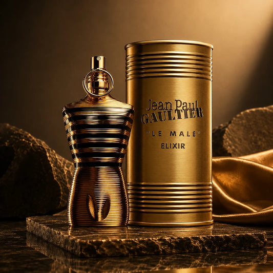Jean Paul Gaultier Le Male Elixir perfume bottle and its cylindrical packaging box, both in a metallic gold finish, displayed on a dark stone pedestal with draped gold fabric in the background.