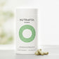 A white cylindrical bottle of Nutrafol Women dietary supplement with a green concentric circle logo, shown on a light surface with two green capsules in front. The background is a soft, blurred white and light green setting.