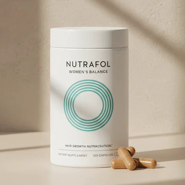 A white cylindrical tub of Nutrafol Women's Balance dietary supplement with a teal concentric circle logo, set on a beige surface with three tan capsules in front. The background is a textured beige wall with soft diagonal shadows.