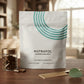 A white stand-up pouch of Nutrafol Women's Balance dietary supplement with a teal curved stripe graphic, displayed on a wooden table next to a small glass jar of brown capsules, a gold comb, and a green leaf. The background is a softly blurred indoor room.