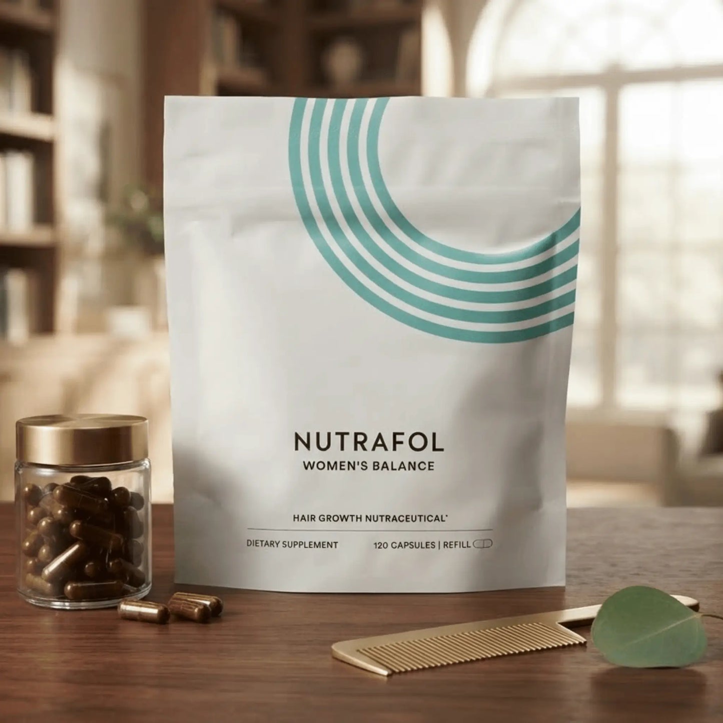 A white stand-up pouch of Nutrafol Women's Balance dietary supplement with a teal curved stripe graphic, displayed on a wooden table next to a small glass jar of brown capsules, a gold comb, and a green leaf. The background is a softly blurred indoor room.
