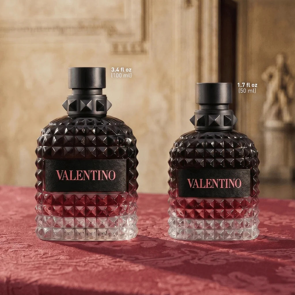 Two Valentino perfume bottles on a red damask fabric surface with a blurred beige interior background. The bottles are faceted dark glass with a red gradient lower half and black caps.