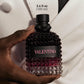 A hand holds a textured Valentino perfume bottle with a black cap and a black label featuring pink lettering, set against a white blazer. Text at the top reads '3.