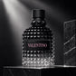 A textured black glass perfume bottle with a faceted diamond pattern, a black cap, and a black label featuring the brand name 'VALENTINO' in pink, displayed on a dark marble pedestal.