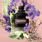 A bottle of Valentino Violet Leaf Eau de Toilette perfume, featuring a textured black bottle with a pink 'VALENTINO' label, surrounded by purple violet flowers and green sage leaves on a soft pink background.