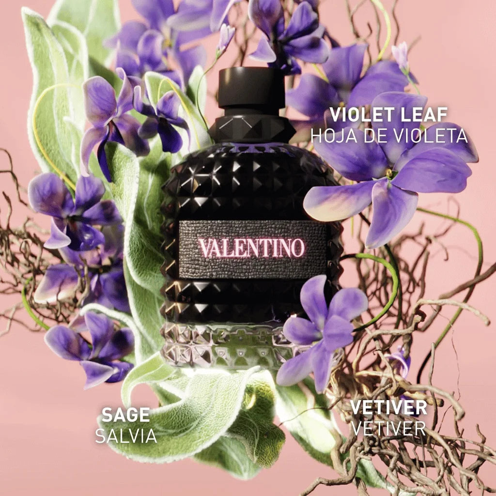 A bottle of Valentino Violet Leaf Eau de Toilette perfume, featuring a textured black bottle with a pink 'VALENTINO' label, surrounded by purple violet flowers and green sage leaves on a soft pink background.