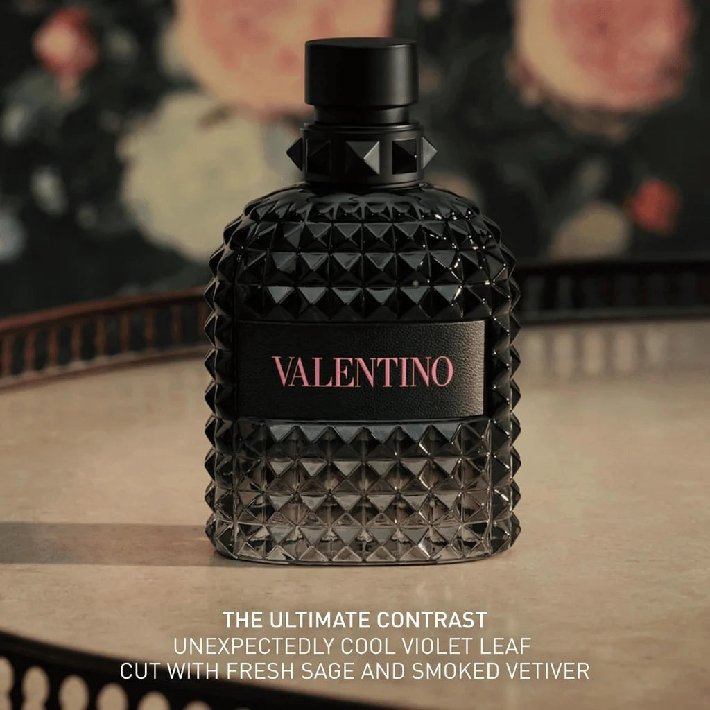 A textured black glass perfume bottle with a black cap and a black label featuring the pink Valentino logo, set on a wooden surface with a blurred floral background. Text at the bottom reads 'THE ULTIMATE CONTRAST UNEXPECTEDLY COOL VIOLET LEAF CUT WITH FRESH SAGE AND SMOKED VETIVER'.