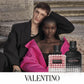 Two models pose on stone steps with Valentino perfume bottles in the foreground. One model wears a black suit jacket, and the other wears a black top with a bright pink shawl.