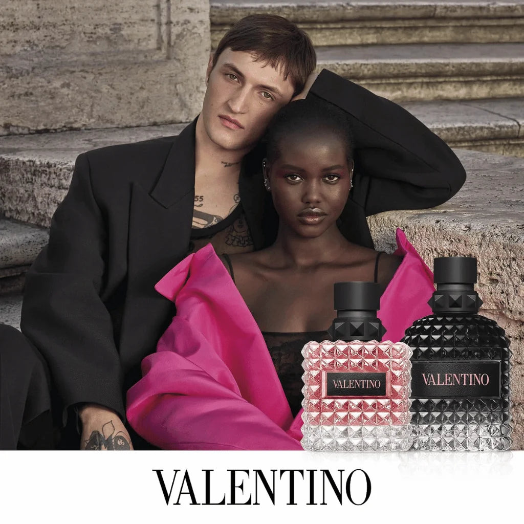 Two models pose on stone steps with Valentino perfume bottles in the foreground. One model wears a black suit jacket, and the other wears a black top with a bright pink shawl.