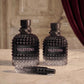 Two Valentino perfume bottles and a travel spray bottle on a marble surface, with a blurred interior background featuring a curtain. The bottles are textured black with pink VALENTINO lettering on the front labels.