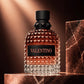 Valentino fragrance bottle with a faceted clear glass body, amber liquid, and a black cap, displayed on a metallic pedestal against a warm brown background with a marble block prop.