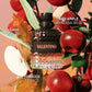 A textured glass perfume bottle labeled VALENTINO with a black cap, surrounded by red apples, green leaves, and autumn leaves on a peach-toned background. White text overlays read SAGE SALVIA, RED APPLE MANZANA ROJA, and TOBACCO TABACO.