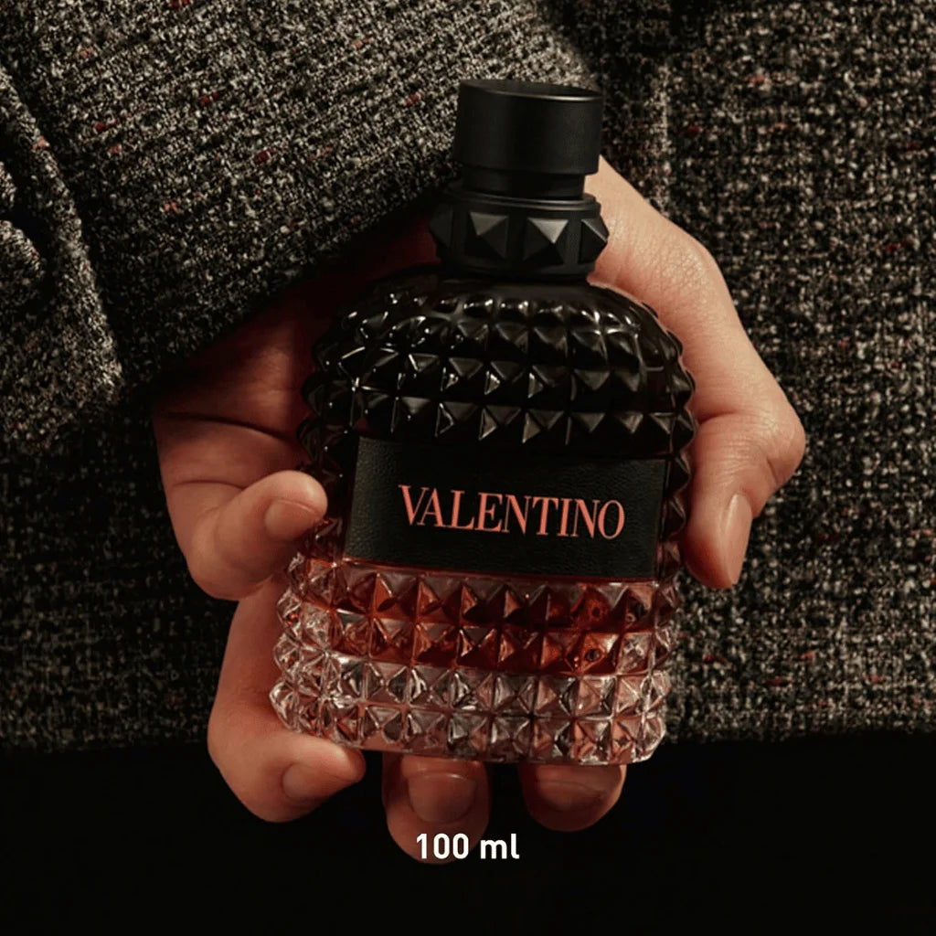 A hand holding a textured Valentino perfume bottle with a black cap and a black label featuring the brand name in red. The bottle has a faceted, diamond-patterned design, with a dark amber liquid in the lower half and a clear upper half.