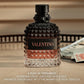 A bottle of Valentino fragrance with a faceted black and clear glass body, a black cap, and a black label with rose-gold lettering, set on a tabletop with blurred background elements and text overlay at the bottom.