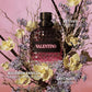 A Valentino perfume bottle with a textured dark glass body and a black cap, surrounded by yellow orchids, lavender flowers, and twigs against a pink background. White text labels read 'VALENTINO', 'VETIVER', 'BOURBON VANILLA', and 'LAVENDER LAVANDA'.