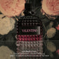 A Valentino perfume bottle with a textured faceted design, featuring a black top and a black label with pink 'VALENTINO' lettering. The bottle is set against a dark floral background with pink and cream roses, with product description text at the bottom.