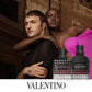 Two models pose in front of a classical building facade, with two Valentino perfume bottles displayed in the lower right foreground. One model wears a sheer black shirt, and the other wears a bright pink draped garment.